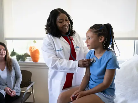 A doctor is examining a young girl with a stethoscope while a woman watches.