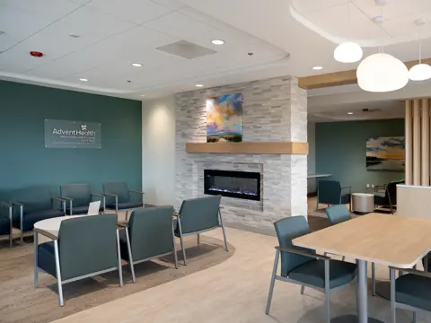 Modern healthcare facility waiting area with teal accent wall featuring AdventHealth Hematology and Oncology signage, comfortable seating arrangements, stone fireplace with wooden mantel, and contemporary pendant lighting