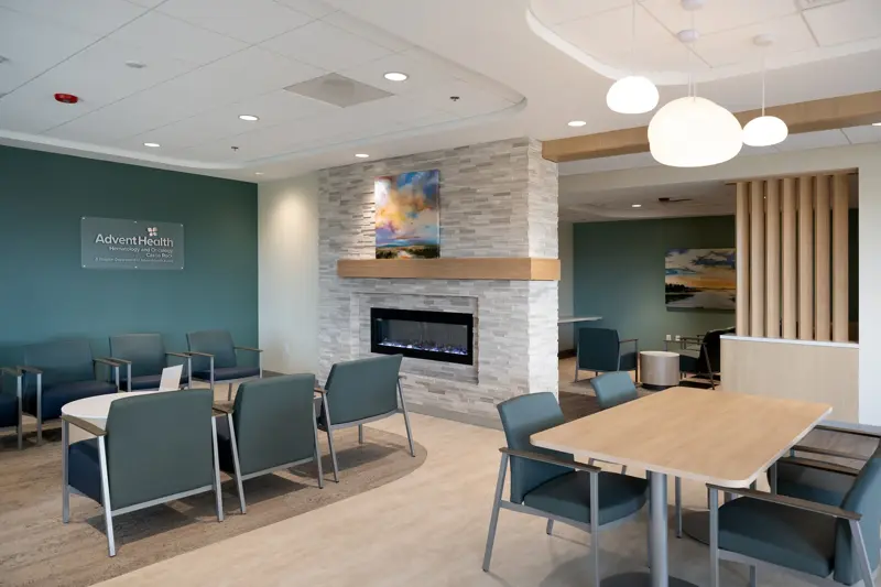 Modern healthcare facility waiting area with teal accent wall featuring AdventHealth Hematology and Oncology signage, comfortable seating arrangements, stone fireplace with wooden mantel, and contemporary pendant lighting