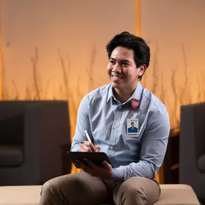 A smiling man in a blue shirt holds a pen and tablet, seated on a bench with two chairs behind him.