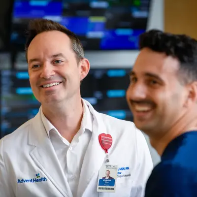 Two health care professionals in scrubs smiling and conversing in a clinical setting with medical monitors visible in the background
