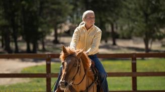 A woman in a yellow shirt and blue jeans rides a brown horse in a fenced pasture.
