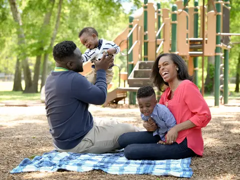 Family of three smiling and playing together in a park with a playground in the background.