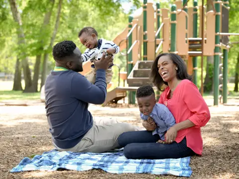 Family of three smiling and playing together in a park with a playground in the background.