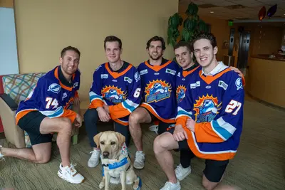Five Orlando Solar Bears hockey players in blue and orange jerseys pose for a group photo with a yellow Labrador service dog in a lobby setting