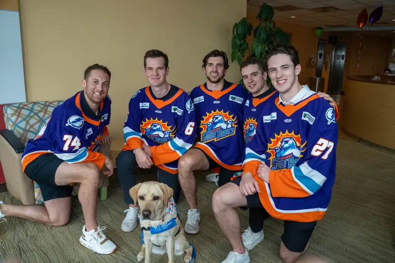 Five Orlando Solar Bears hockey players in blue and orange jerseys pose for a group photo with a yellow Labrador service dog in a lobby setting