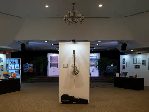 Art gallery with paintings and framed works lining the walls, centered on a displayed guitar and case.