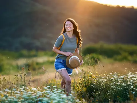 Happy woman is walking on the grass, holding a hat in her hand, while the sun sets behind the mountain.