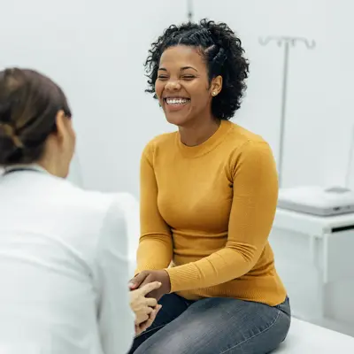 Woman in yellow sweater smiling while sitting on an examination table and speaking with her primary care provider in a bright medical office