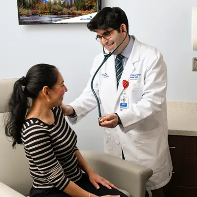 Medical resident in white coat using stethoscope to examine smiling female patient seated in exam room