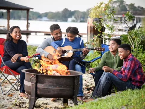 Family gathered around a fire pit, with one member playing a guitar and others roasting marshmallows.