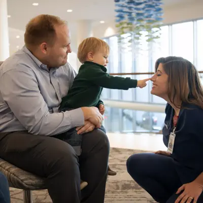 A father holds a child while a nurse interacts with them in a hospital setting.