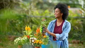 A woman in a denim jacket and gloves holds a potted plant, smiling at the camera with various flowers in the background.