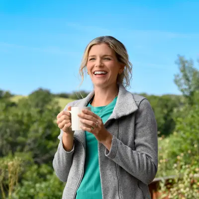 A smiling woman in a green shirt and gray jacket holds a white cup outdoors.