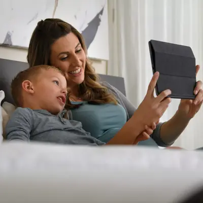 A mother and son are lying on a bed, smiling and looking at a tablet.