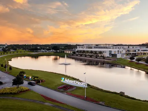 Aerial view of AdventHealth Sebring facility at dusk with a pond and fountain in the foreground, surrounded by landscaped grounds under a vibrant orange and yellow sunset sky