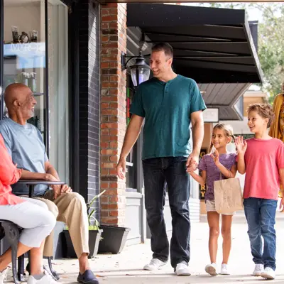 A family of four walks down a sidewalk, smiling and waving at two seated individuals outside a store.