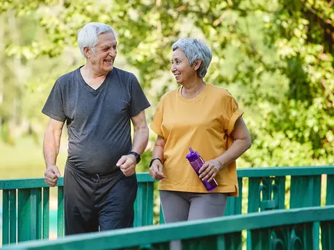 An older couple in athletic wear walking together on a green bridge outdoors, smiling and enjoying each other's company during their exercise routine