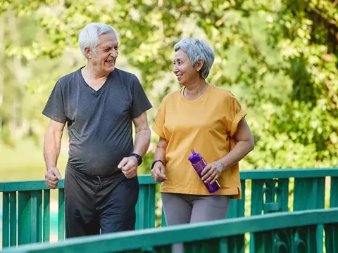 An older couple in athletic wear walking together on a green bridge in a sunny park, smiling and enjoying outdoor exercise