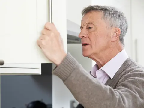 Senior man in a gray sweater opening a white kitchen cabinet in a modern home