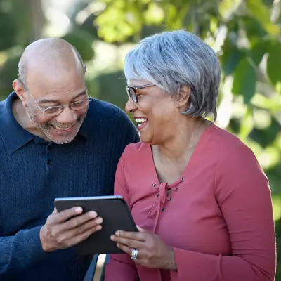 Older couple smiling and looking at a tablet together while sitting on a park bench outdoors