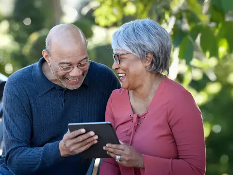 Older couple smiling and looking at a tablet together while sitting on a park bench outdoors