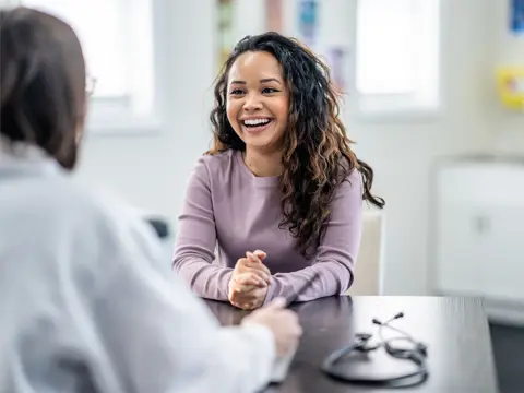 Smiling woman in purple sweater sitting at a desk during a consultation with a healthcare provider in a bright medical office