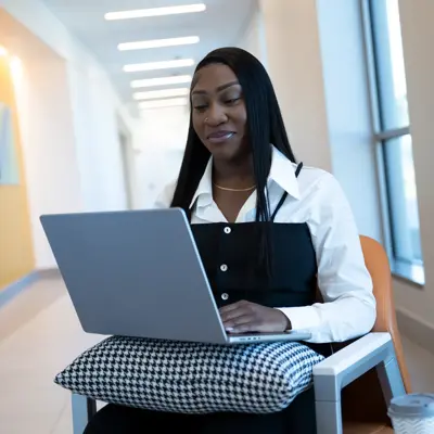 A woman in a white shirt and black vest works on a laptop in a hallway.