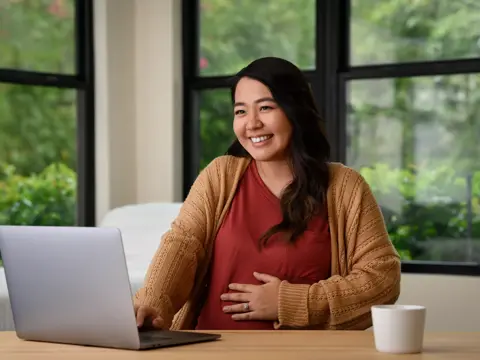 A smiling woman at a desk with a laptop, hand on her belly, and a cup nearby.