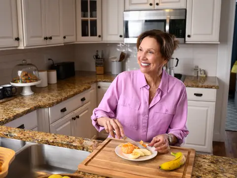 A smiling woman in a pink shirt is cutting fruit on a cutting board in a kitchen.