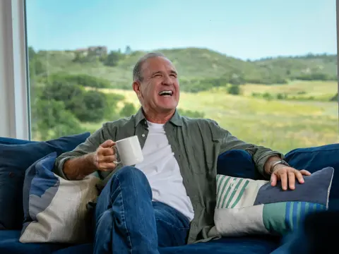Man sitting on a couch, holding a coffee mug, smiling at the camera with a scenic view outside the window.