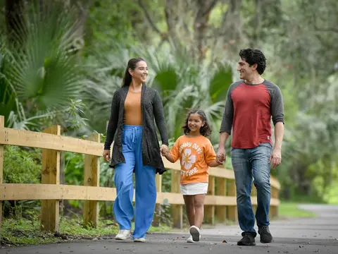 A smiling family of three walks down a park path, holding hands.