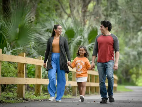 A smiling family of three walks down a park path, holding hands.