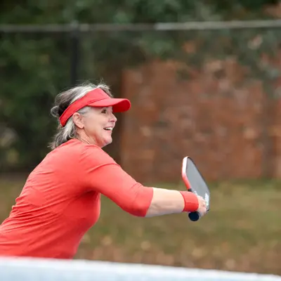 A woman in a red shirt and visor plays pickleball, hitting a yellow ball with a paddle.
