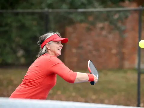 A woman in a red shirt and visor plays pickleball, hitting a yellow ball with a paddle.
