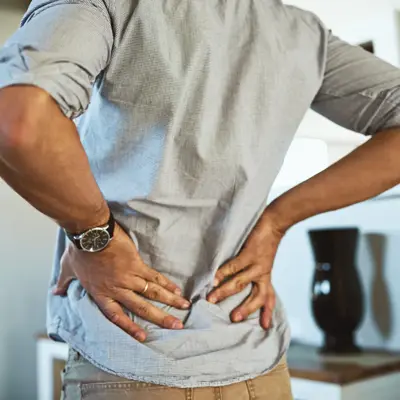 Person standing indoors with both hands pressed against their lower back, suggesting lower back discomfort.