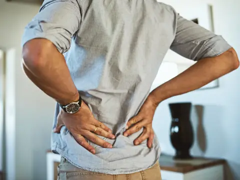 Person standing indoors with both hands pressed against their lower back, suggesting lower back discomfort.