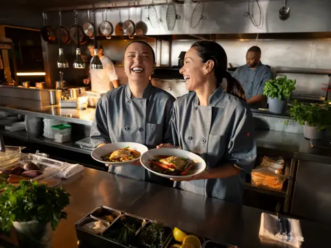 Two chefs in a kitchen, smiling and holding plates of food.