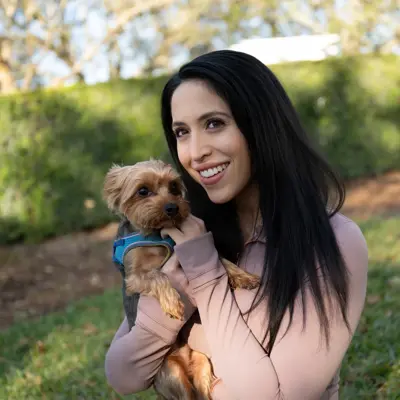 A smiling woman with long black hair holds a small brown dog in a park.