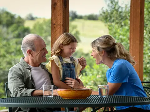 Grandfather, mother, and daughter sitting at a table, smiling and conversing.