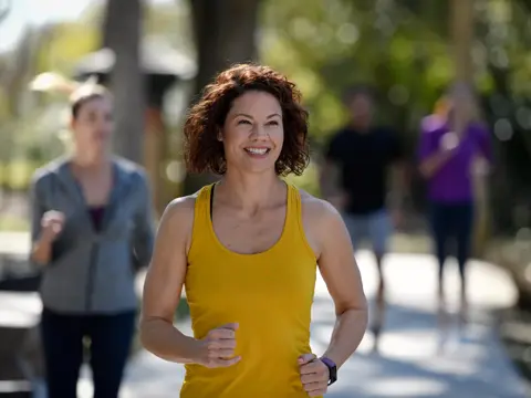 A woman in a yellow tank top jogs with a smile, leading a group of runners on a sunny day.