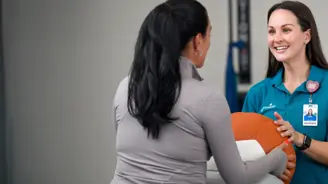 Two women stand facing each other, smiling, while holding a large orange and white exercise ball.