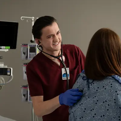 A smiling male nurse with a stethoscope around his neck examines a female patient in a hospital room.