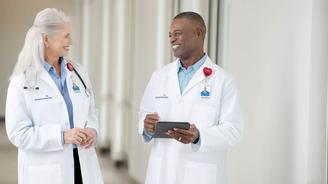 Two AdventHealth doctors in white coats, smiling and conversing in a hospital corridor.