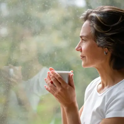 A Middle-aged woman drinking from a cup, looking out a window.