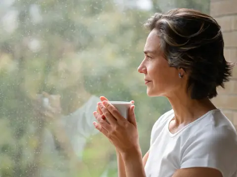 A Middle-aged woman drinking from a cup, looking out a window.