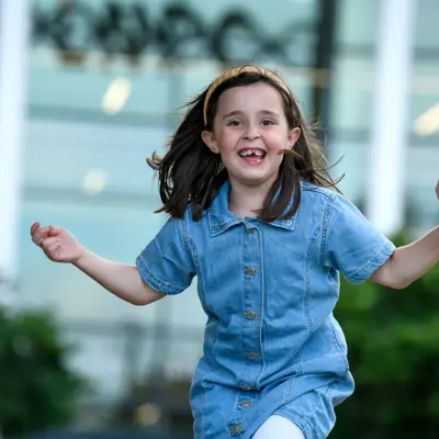 A young girl in a blue denim outfit joyfully jumps in the air with a wide smile.