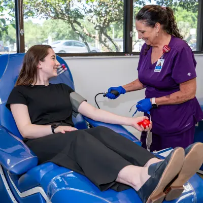 A woman is seated in a blue chair while a nurse checks her blood pressure.