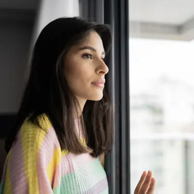 Woman in a colorful sweater looking thoughtfully out a window with natural light, conveying a contemplative and emotional moment