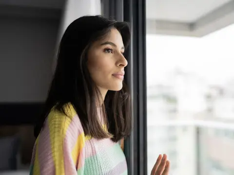 Woman in a colorful sweater looking thoughtfully out a window with natural light, conveying a contemplative and emotional moment